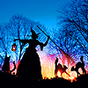 witchofthebough: A witch wields her broom defensively up in the air with her left hand and carries a lantern in her right as she travels the woods with her cats against a bright orange and royal blue sunset. (Light Beyond)