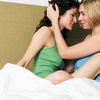 bluflamingo: two white girls, one dark hair, one blonde, sitting up on bed. blonde has her hand on brunette's face (two girls kissing)